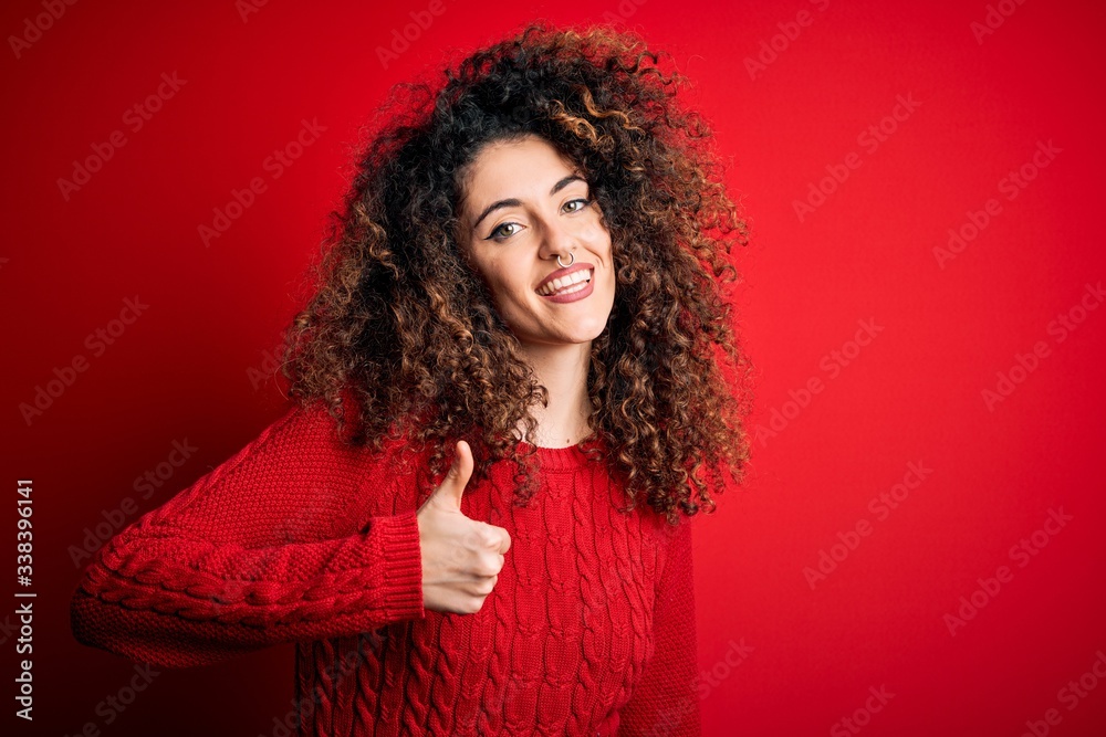 Young beautiful woman with curly hair and piercing wearing casual red sweater doing happy thumbs up gesture with hand. Approving expression looking at the camera showing success. Young beautiful woman with curly hair and piercing wearing casual red sweater doing happy thumbs up gesture with hand. Approving expression looking at the camera showing success.
