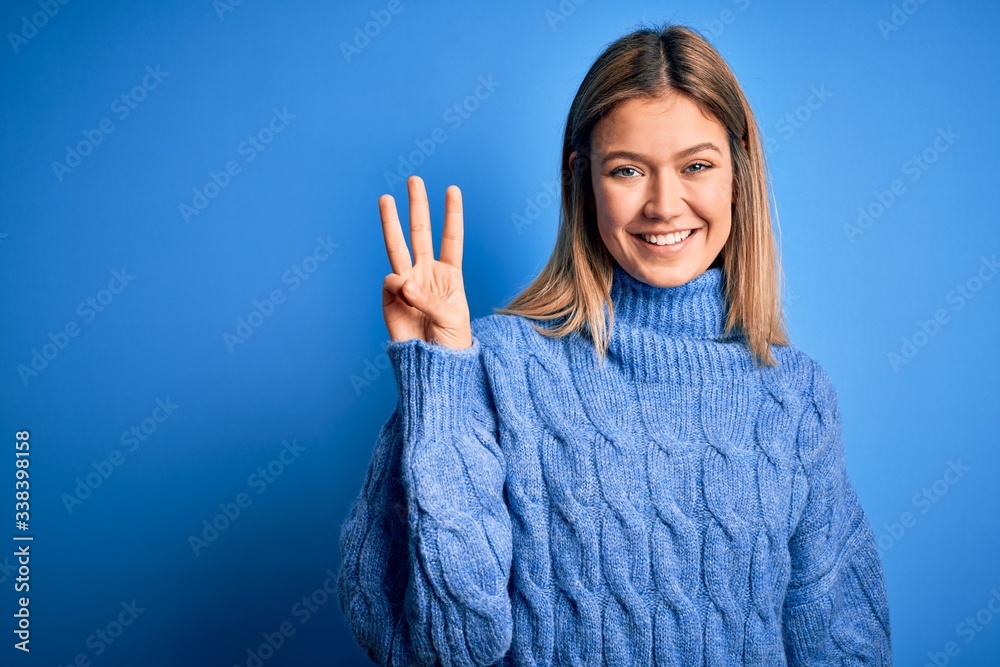 Young beautiful blonde woman wearing winter wool sweater over blue isolated background showing and pointing up with fingers number three while smiling confident and happy. Young beautiful blonde woman wearing winter wool sweater over blue isolated background showing and pointing up with fingers number three while smiling confident and happy.