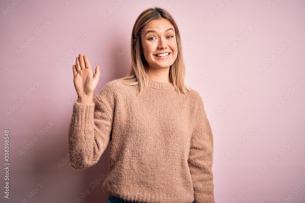 Young beautiful blonde woman wearing winter wool sweater over pink isolated background Waiving saying hello happy and smiling, friendly welcome gesture