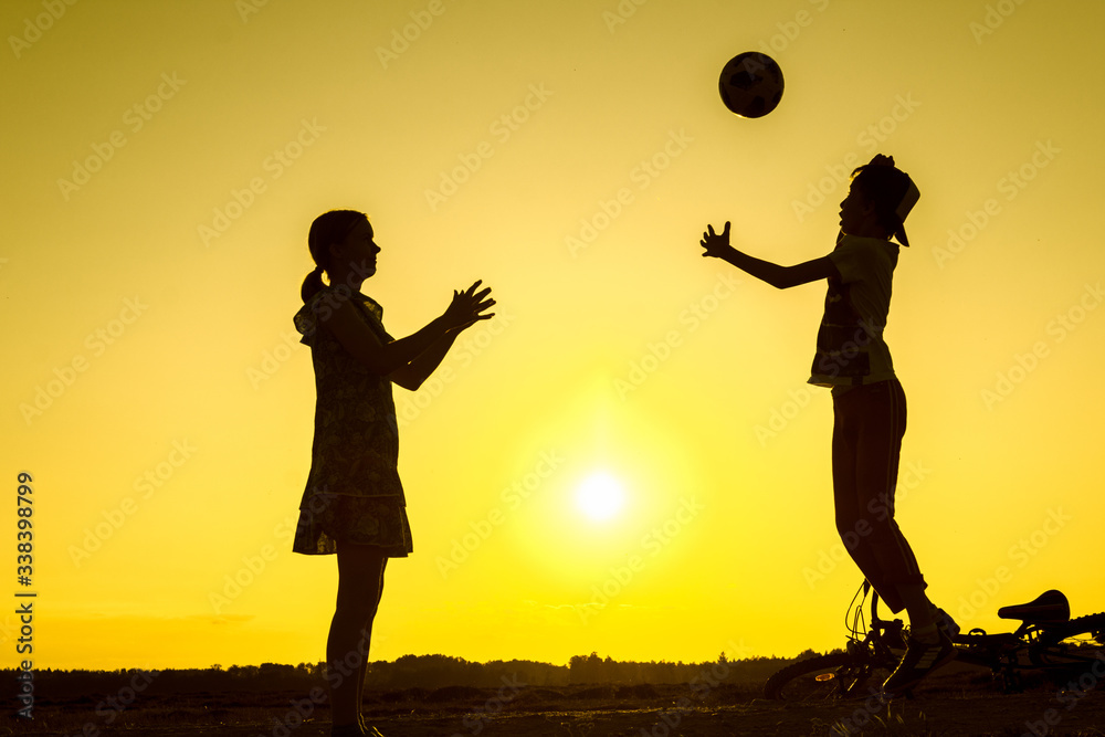Brother with sister are playing with ball in nature, bicycle lies nearby, silhouettes of playing children at sunset in countryside Brother with sister are playing with ball in nature, bicycle lies nearby, silhouettes of playing children at sunset in countryside
