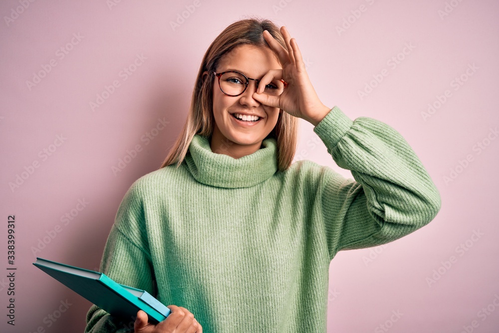 Young beautiful woman wearing glasses holding books over isolated pink background with happy face smiling doing ok sign with hand on eye looking through fingers Young beautiful woman wearing glasses holding books over isolated pink background with happy face smiling doing ok sign with hand on eye looking through fingers