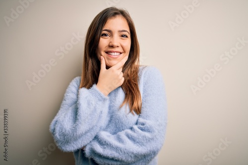 Beautiful young woman wearing casual winter sweater standing over isolated background looking confident at the camera smiling with crossed arms and hand raised on chin. Thinking positive. © Krakenimages.com