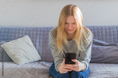 Front view of a sad blonde woman checking phone sitting sitting on the sofa in the living room at home. Technology concept and digital devices. Female blogger chatting with friends in social network.