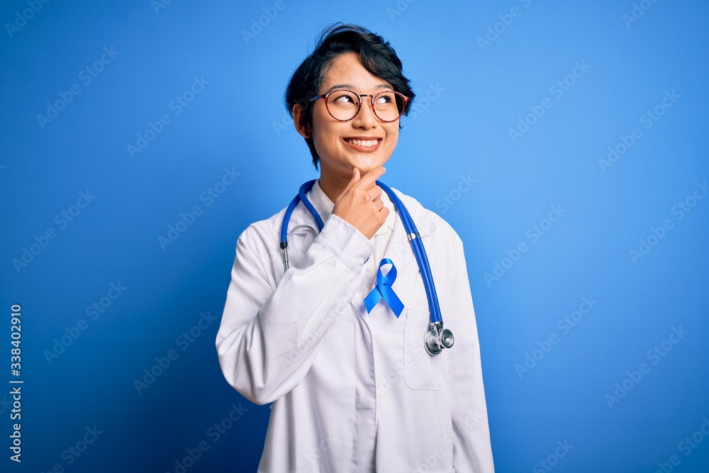 Young beautiful asian doctor girl wearing stethoscope and coat with blue cancer ribbon with hand on chin thinking about question, pensive expression. Smiling with thoughtful face. Doubt concept. Young beautiful asian doctor girl wearing stethoscope and coat with blue cancer ribbon with hand on chin thinking about question, pensive expression. Smiling with thoughtful face. Doubt concept.