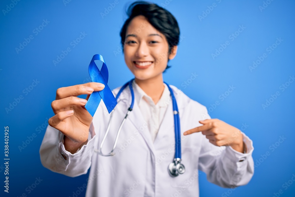 Young beautiful asian doctor girl wearing stethoscope and coat holding blue cancer ribbon with surprise face pointing finger to himself Young beautiful asian doctor girl wearing stethoscope and coat holding blue cancer ribbon with surprise face pointing finger to himself