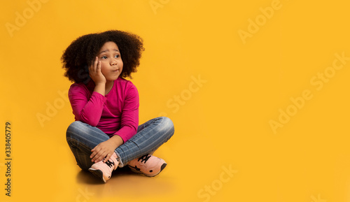 Pensive little girl sitting on floor over yellow background and thinking
