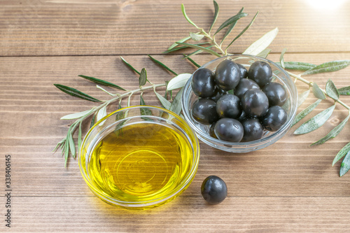 Olives and olive oil in glass transparent bowl , branches of olive tree on the wooden background in sunlights.