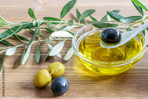 Olive oil in the glass transparent bowl, spoon with black olive , olive tree branches on the wooden background closeup. Mediterranean healthy diet food.