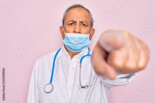 Senior hoary doctor man wearing medical mask and stethoscope over pink background pointing with finger to the camera and to you, hand sign, positive and confident gesture from the front