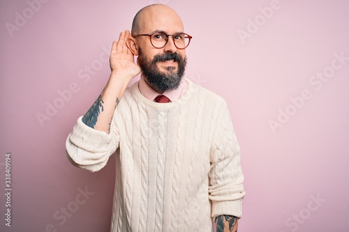 Handsome bald man with beard and tattoo wearing glasses and sweater over pink background smiling with hand over ear listening an hearing to rumor or gossip. Deafness concept.