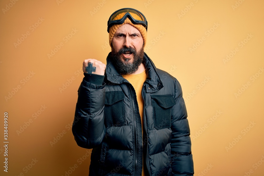 Handsome skier bald man with beard skiing wearing snow sportswear and ski goggles angry and mad raising fist frustrated and furious while shouting with anger. Rage and aggressive concept. Handsome skier bald man with beard skiing wearing snow sportswear and ski goggles angry and mad raising fist frustrated and furious while shouting with anger. Rage and aggressive concept.