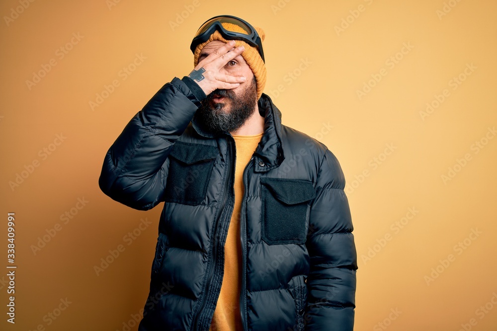 Handsome skier bald man with beard skiing wearing snow sportswear and ski goggles peeking in shock covering face and eyes with hand, looking through fingers with embarrassed expression. Handsome skier bald man with beard skiing wearing snow sportswear and ski goggles peeking in shock covering face and eyes with hand, looking through fingers with embarrassed expression.
