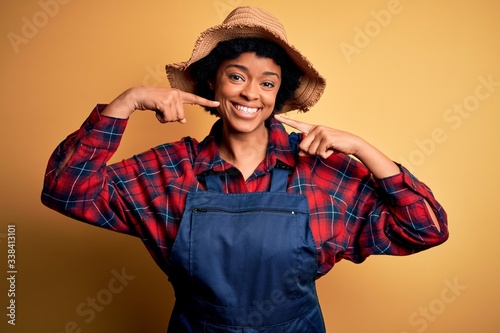 Young African American afro farmer woman with curly hair wearing apron and hat smiling cheerful showing and pointing with fingers teeth and mouth. Dental health concept.