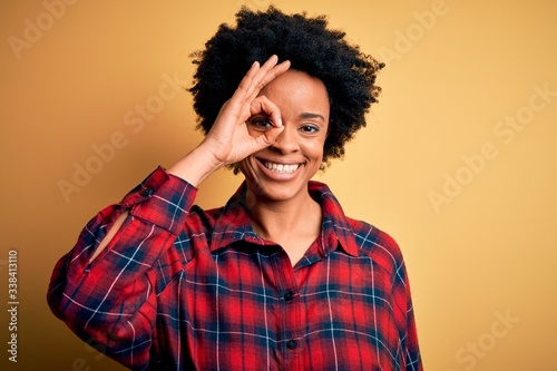 Young beautiful African American afro woman with curly hair wearing casual shirt doing ok gesture with hand smiling, eye looking through fingers with happy face. © Krakenimages.com