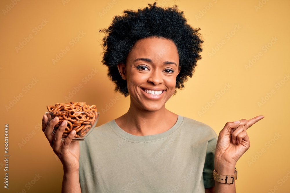 Young African American afro woman with curly hair holding bowl with baked German pretzels very happy pointing with hand and finger to the side Young African American afro woman with curly hair holding bowl with baked German pretzels very happy pointing with hand and finger to the side
