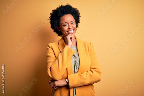 Young beautiful African American afro businesswoman with curly hair wearing yellow jacket looking confident at the camera with smile with crossed arms and hand raised on chin. Thinking positive. © Krakenimages.com
