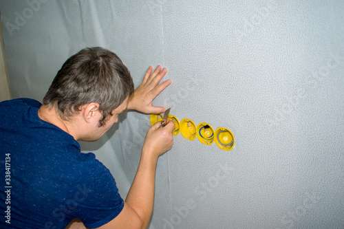 The man cuts the wallpaper on the walls in the places of the sockets with a knife.