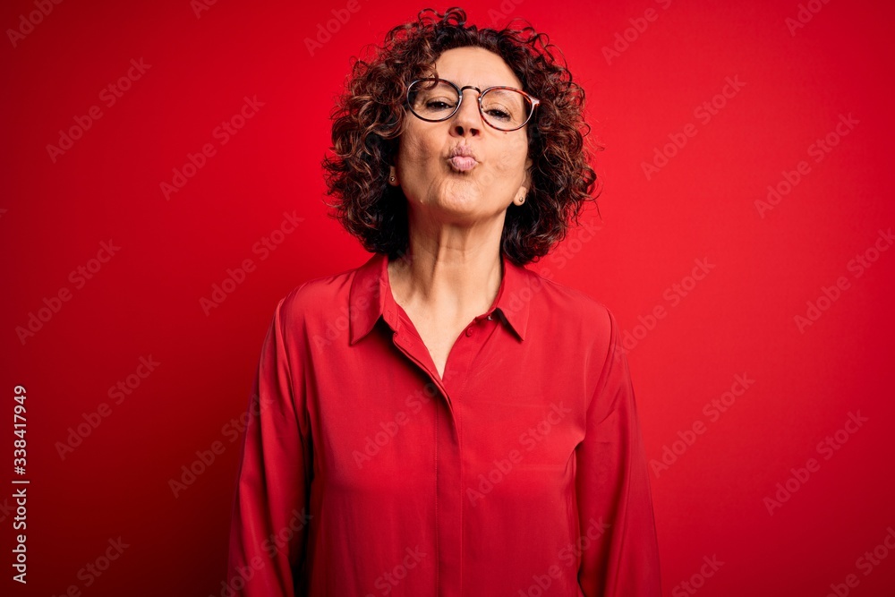 Middle age beautiful curly hair woman wearing casual shirt and glasses over red background looking at the camera blowing a kiss on air being lovely and sexy. Love expression. Middle age beautiful curly hair woman wearing casual shirt and glasses over red background looking at the camera blowing a kiss on air being lovely and sexy. Love expression.