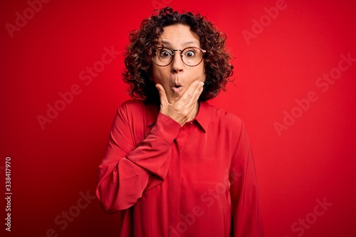 Middle age beautiful curly hair woman wearing casual shirt and glasses over red background Looking fascinated with disbelief, surprise and amazed expression with hands on chin