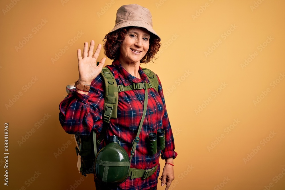 Middle age curly hair hiker woman hiking wearing backpack and water canteen using binoculars Waiving saying hello happy and smiling, friendly welcome gesture