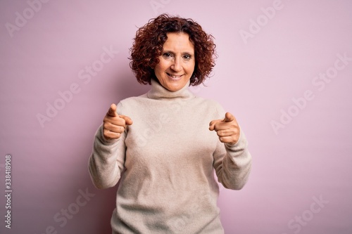 Middle age beautiful curly hair woman wearing casual turtleneck sweater over pink background pointing fingers to camera with happy and funny face. Good energy and vibes.