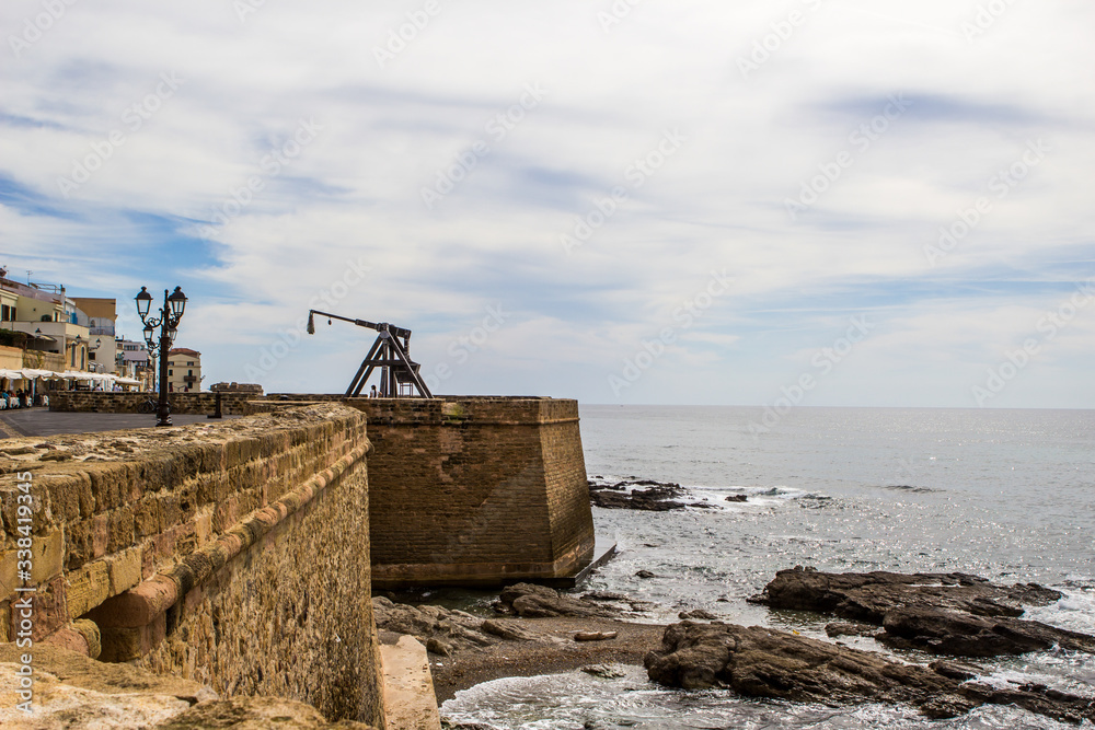 Grey rocks, brick wall with ancient catapult, waves with foam and sky with clouds in Alghero, Sardinia, Italia. Beautiful view on the seafront Alghero.