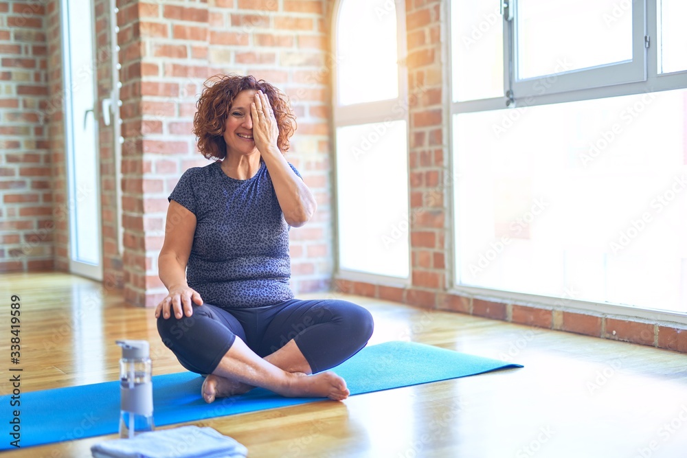 Middle age beautiful sportswoman wearing sportswear sitting on mat practicing yoga at home covering one eye with hand, confident smile on face and surprise emotion. Middle age beautiful sportswoman wearing sportswear sitting on mat practicing yoga at home covering one eye with hand, confident smile on face and surprise emotion.