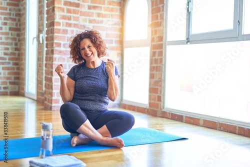 Middle age beautiful sportswoman wearing sportswear sitting on mat practicing yoga at home very happy and excited doing winner gesture with arms raised, smiling and screaming for success. Celebration