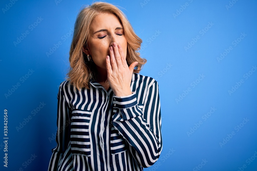 Middle age beautiful blonde woman wearing casual striped shirt standing over blue background bored yawning tired covering mouth with hand. Restless and sleepiness.