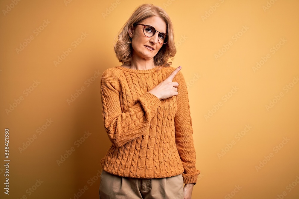 Middle age beautiful blonde woman wearing casual sweater and glasses over yellow background Pointing with hand finger to the side showing advertisement, serious and calm face Middle age beautiful blonde woman wearing casual sweater and glasses over yellow background Pointing with hand finger to the side showing advertisement, serious and calm face