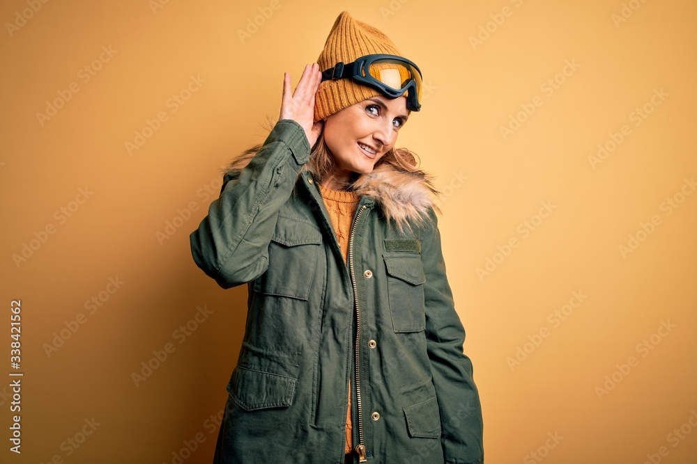 Middle age beautiful blonde skier woman wearing snow sportwear and ski goggles smiling with hand over ear listening an hearing to rumor or gossip. Deafness concept.