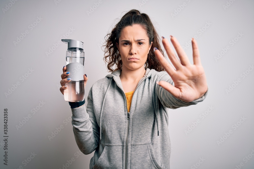 Beautiful woman with curly hair doing sport drinking bottle of water over white background with open hand doing stop sign with serious and confident expression, defense gesture Beautiful woman with curly hair doing sport drinking bottle of water over white background with open hand doing stop sign with serious and confident expression, defense gesture