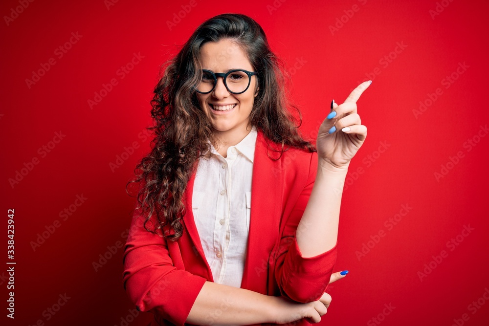 Young beautiful woman with curly hair wearing jacket and glasses over red background with a big smile on face, pointing with hand and finger to the side looking at the camera. Young beautiful woman with curly hair wearing jacket and glasses over red background with a big smile on face, pointing with hand and finger to the side looking at the camera.