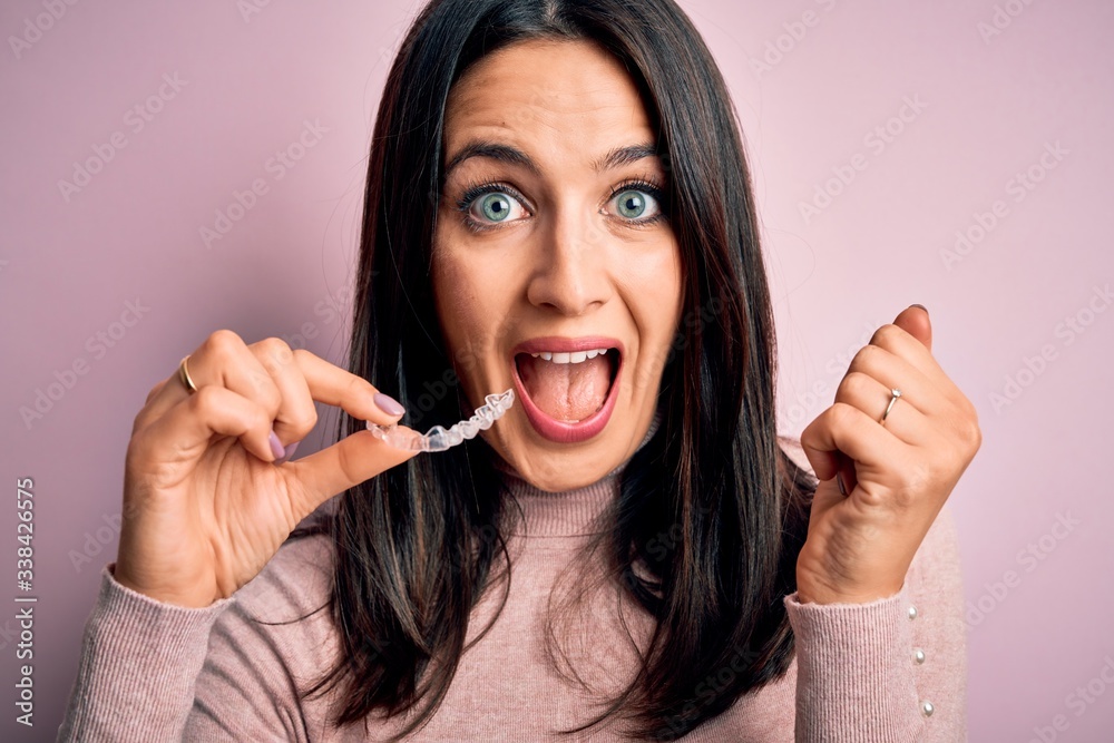 Young woman with blue eyes holding clear aligner standing over pink background screaming proud and celebrating victory and success very excited, cheering emotion Young woman with blue eyes holding clear aligner standing over pink background screaming proud and celebrating victory and success very excited, cheering emotion