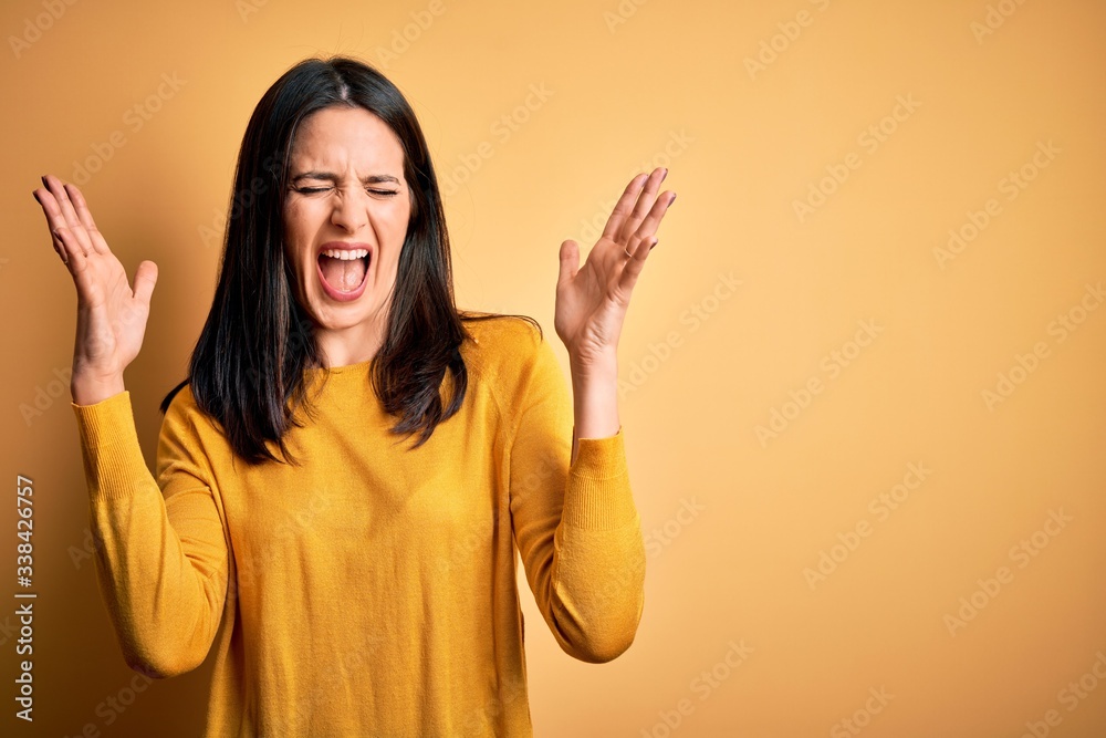 Young brunette woman with blue eyes wearing casual sweater over yellow background celebrating mad and crazy for success with arms raised and closed eyes screaming excited. Winner concept