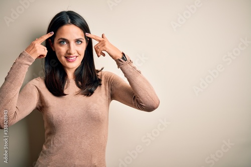 Young brunette woman with blue eyes wearing casual sweater over isolated white background smiling pointing to head with both hands finger, great idea or thought, good memory
