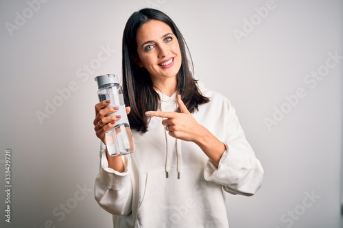 Young brunette sporty woman with blue eyes holding water bottle over white background very happy pointing with hand and finger