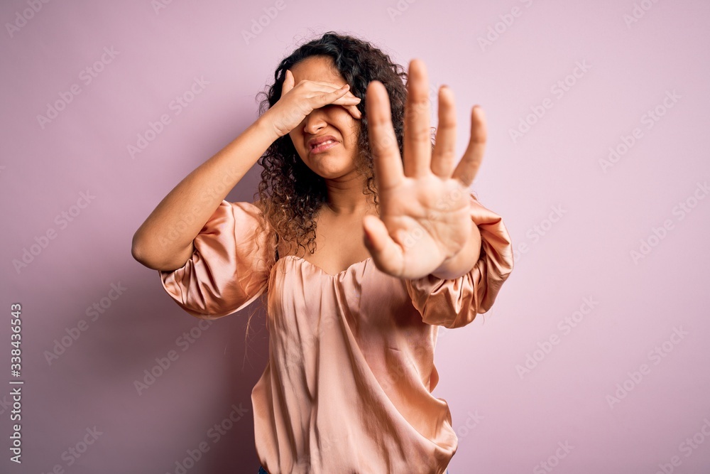 Young beautiful woman with curly hair wearing casual t-shirt standing over pink background covering eyes with hands and doing stop gesture with sad and fear expression. Embarrassed and negative