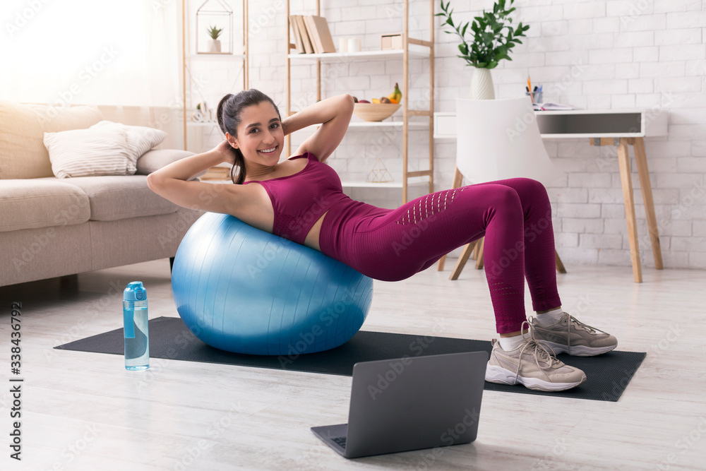 Stay home, stay active. Hispanic woman working out with fitness ball near laptop in living room Stay home, stay active. Hispanic woman working out with fitness ball near laptop in living room