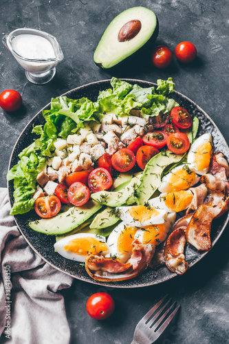 Cobb salad with chicken, avocado, tomatoes, eggs, bacon and cheese with sauce on a black table close up.