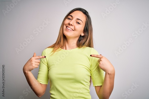 Beautiful blonde woman with blue eyes wearing green casual t-shirt over white background looking confident with smile on face, pointing oneself with fingers proud and happy.