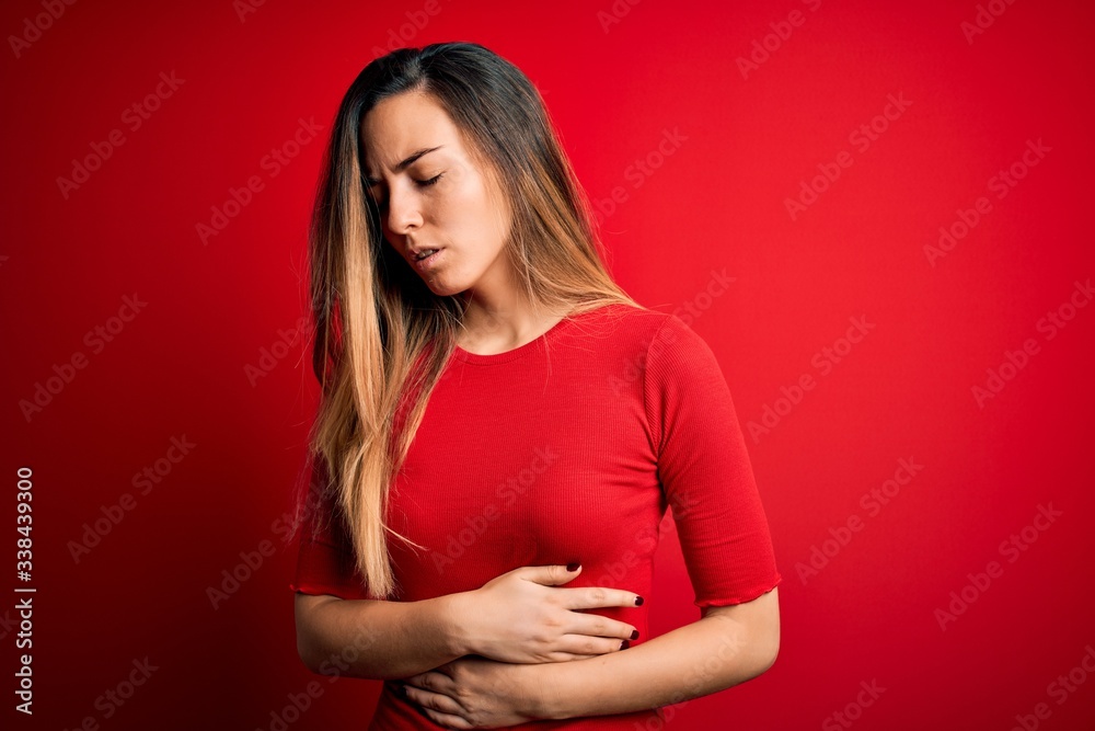 Young beautiful blonde woman with blue eyes wearing casual t-shirt over red background with hand on stomach because indigestion, painful illness feeling unwell. Ache concept. Young beautiful blonde woman with blue eyes wearing casual t-shirt over red background with hand on stomach because indigestion, painful illness feeling unwell. Ache concept.