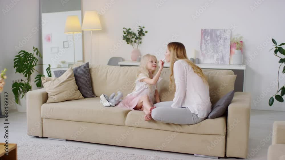 Tracking of cute girl with blond hair sitting on couch and putting lipstick on her mother Tracking of cute girl with blond hair sitting on couch and putting lipstick on her mother