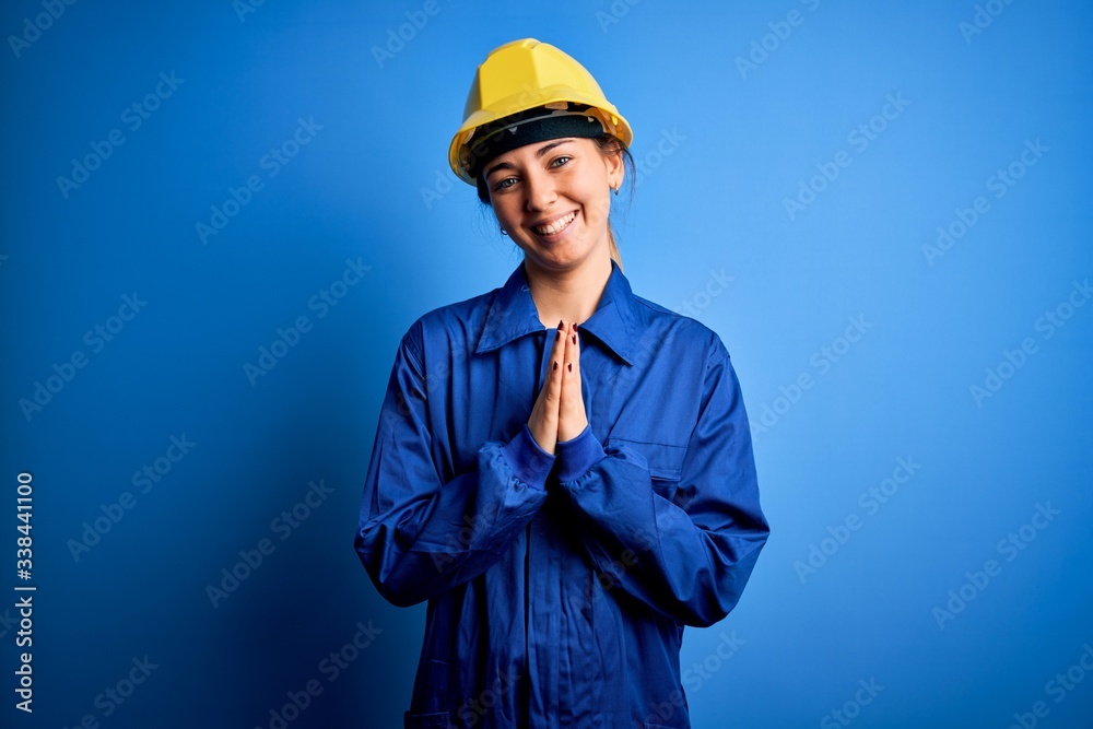 Young beautiful worker woman with blue eyes wearing security helmet and uniform praying with hands together asking for forgiveness smiling confident. Young beautiful worker woman with blue eyes wearing security helmet and uniform praying with hands together asking for forgiveness smiling confident.