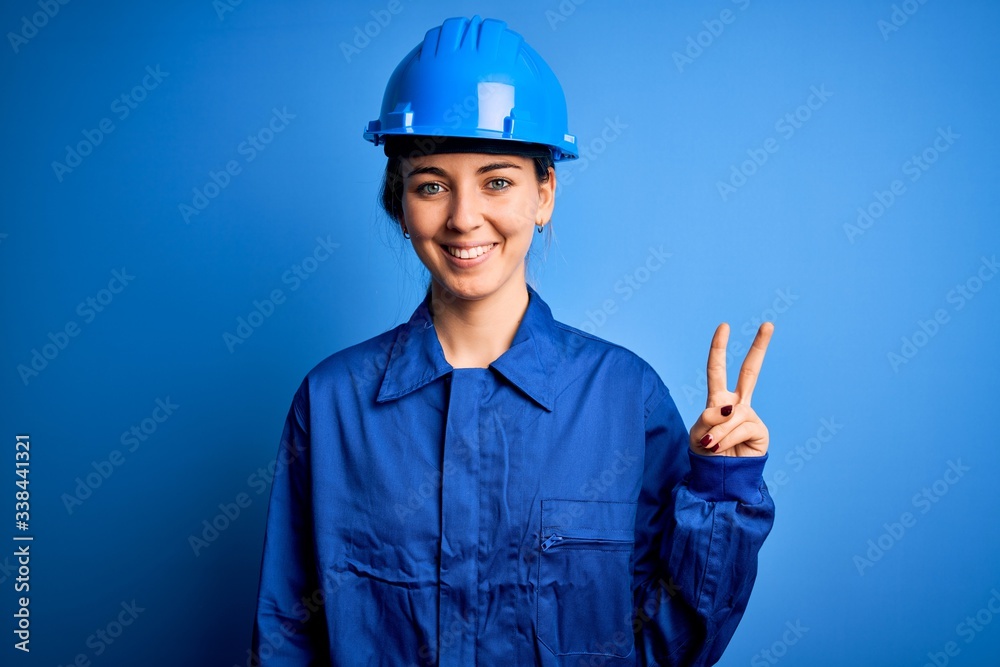 Young beautiful worker woman with blue eyes wearing security helmet and uniform smiling with happy face winking at the camera doing victory sign. Number two. Young beautiful worker woman with blue eyes wearing security helmet and uniform smiling with happy face winking at the camera doing victory sign. Number two.