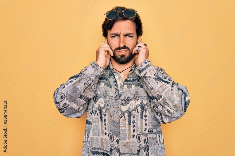 Young handsome hispanic bohemian man wearing hippie style and sunglasses covering ears with fingers with annoyed expression for the noise of loud music. Deaf concept. Young handsome hispanic bohemian man wearing hippie style and sunglasses covering ears with fingers with annoyed expression for the noise of loud music. Deaf concept.