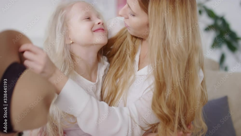 Slowmo of happy mother sitting on couch and hugging adorable little girl. She is taking off wide brim hat from her head and kissing her forehead, then they are looking at camera and smiling Slowmo of happy mother sitting on couch and hugging adorable little girl. She is taking off wide brim hat from her head and kissing her forehead, then they are looking at camera and smiling