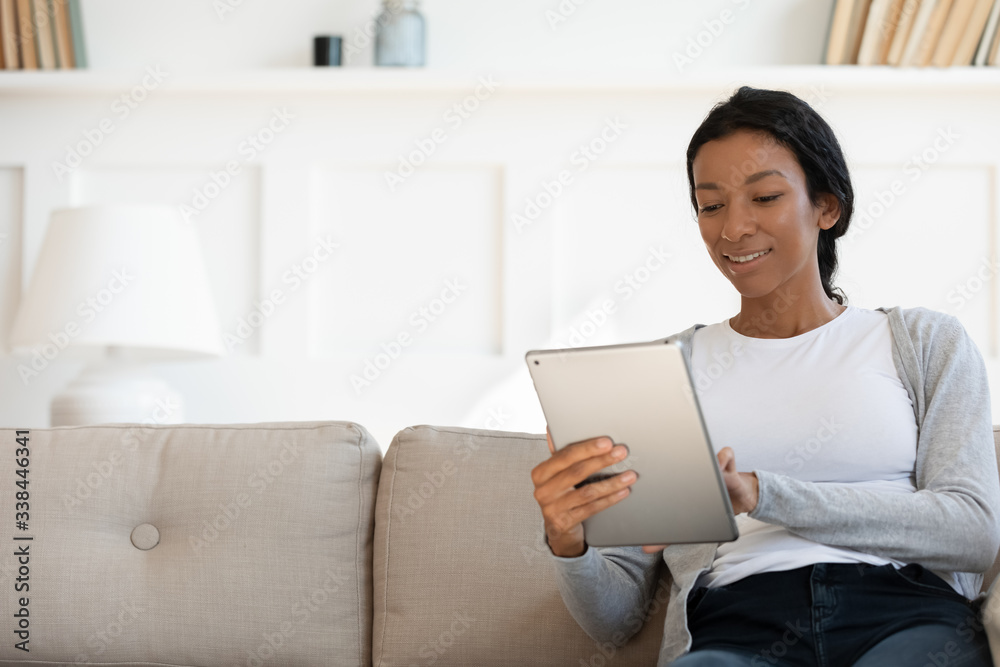 Smiling african american young woman hold tablet and sit on couch in modern living room. Shopping online from home in online store, watching video, surfing or browsing internet. Image with copy space Smiling african american young woman hold tablet and sit on couch in modern living room. Shopping online from home in online store, watching video, surfing or browsing internet. Image with copy space