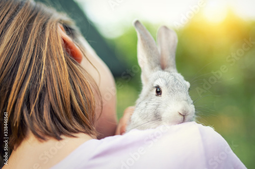 Cute adorable sweet little rabbit sitting on shoulder of young adult brunette woman at warm sunset time country outdoors. Animal friendship and care concept