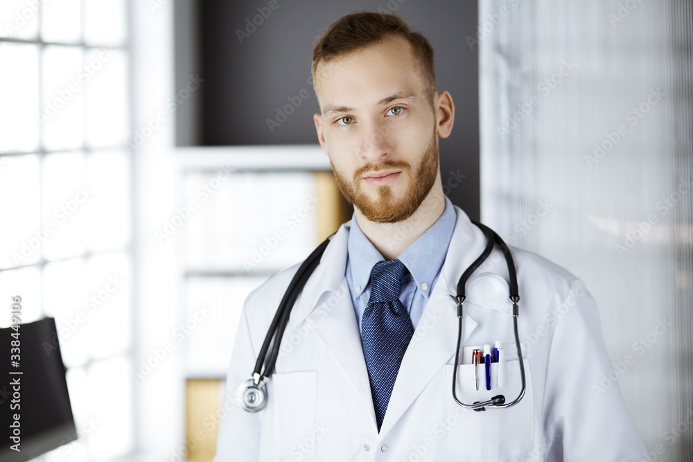 Red-bearded doctor standing straight in clinic near his working place. Portrait of physician. Medicine and healthcare concept